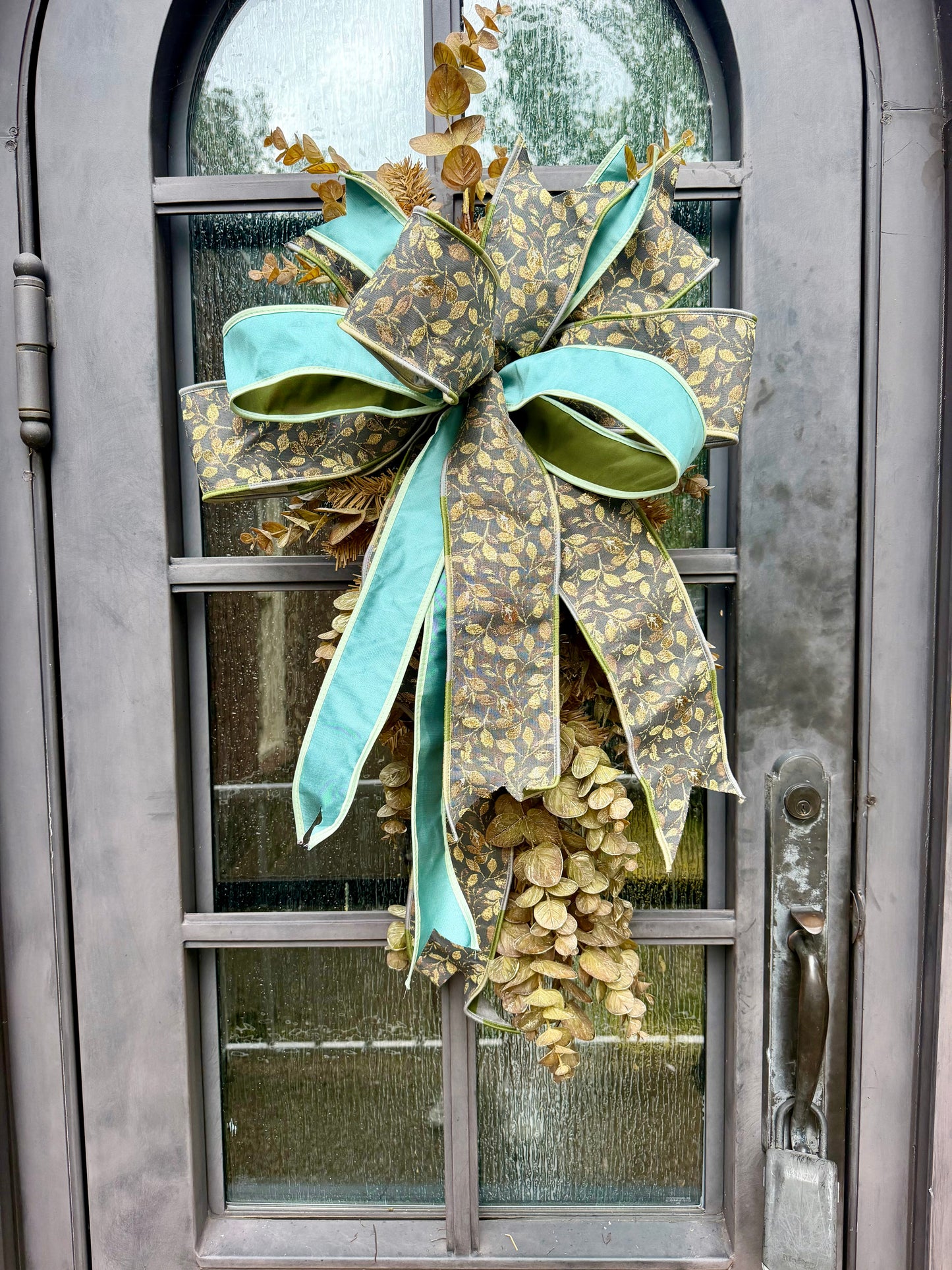 Handmade JME fall door swag made with faux brown eucalyptus. Paired with a beautiful floral grey and teal JME bow. About 3 feet long by 21 inches wide. *Some fluffing may be required upon receiving.