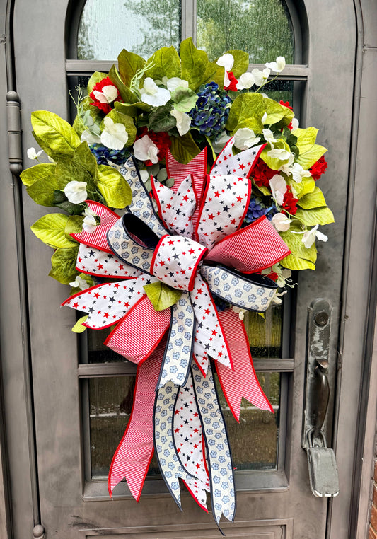 JME handmade faux wreath of magnolias white sweetpeas, blue hydrangeas, and red geraniums with mixed greenery. Paired with a JME patriotic star bow! About 27 inches in diameter.