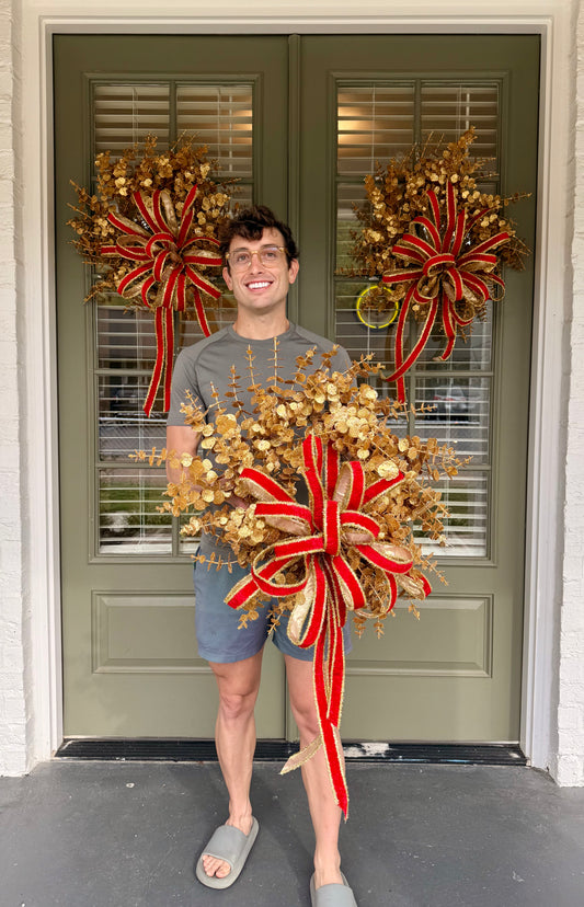 Glittered gold faux eucalyptus. Tied with a JME red velvet and gold tinsel bow. About 20 inches wide.